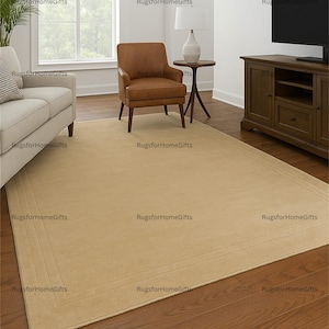 May include: A beige rectangular area rug with a subtle border detail, placed on a dark wood floor. The rug is in a living room setting, with a sofa, armchair, and TV cabinet visible.