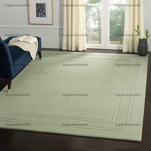 Könnte beinhalten: Ein rechteckiger, hellgrüner Teppich mit einem Bordürendesign. Der Teppich liegt auf einem dunklen Holzboden, mit einem blauen Sofa und einem Fenster mit weißen Vorhängen im Hintergrund. Der Teppich ist ein Einrichtungsgegenstand.