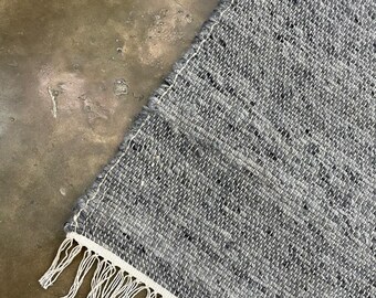 May include: A section of a woven rug with a grey and white speckled pattern. The rug features a white border and white fringe along one edge. The rug is made of a durable material.