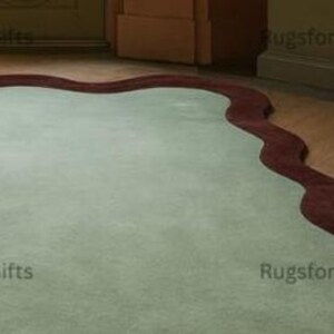 May include: A pale green rug with a dark red, wavy border. The rug has a rectangular shape and a soft, plush texture. The design is simple and elegant, suitable for a living room or bedroom.