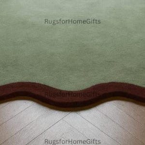 May include: A sage green rug with a wavy, dark red border. The rug is placed on a wooden floor with a herringbone pattern. The rug's unique shape and colour palette add a touch of elegance to any room. The text "RugsforHomeGifts" is visible in the corners.