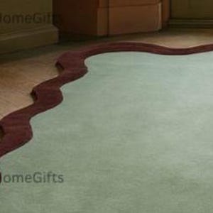 May include: A pale green rug with a distinctive, undulating burgundy edge. The rug is positioned on a wooden floor, offering a contrast in textures and hues. The design is contemporary, adding a touch of sophistication to a room.
