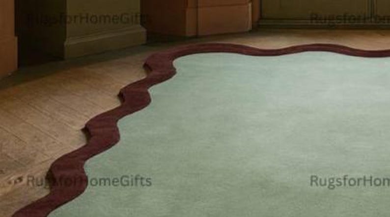 May include: A pale green rug with a distinctive, undulating burgundy edge. The rug is positioned on a wooden floor, offering a contrast in textures and hues. The design is contemporary, adding a touch of sophistication to a room.