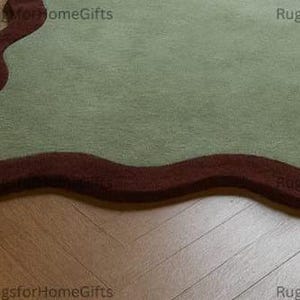 May include: A sage green rug with a dark brown, wavy border. The rug has a soft, textured appearance and an organic shape. The rug is placed on a wooden floor.