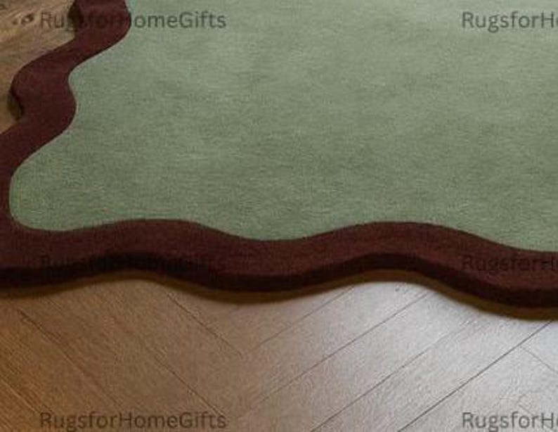 May include: A sage green rug with a dark brown, wavy border. The rug has a soft, textured appearance and an organic shape. The rug is placed on a wooden floor.