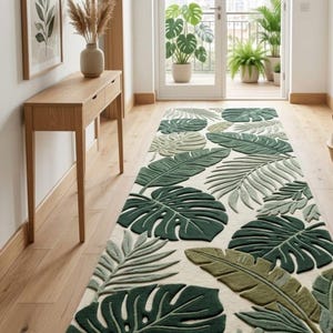 Könnte beinhalten: Ein langer, rechteckiger Teppich mit einem sich wiederholenden Muster aus großen tropischen Monstera- und Palmenblättern in verschiedenen Schattierungen von Dunkelgrün, Oliv und Salbei auf cremefarbenem Grund mit getufteter Struktur.