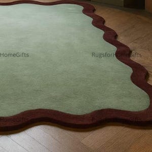 May include: A sage green rug with a wavy, dark red border. The rug has an irregular shape. The rug is placed on a wooden floor, adding a touch of colour and texture to the room.