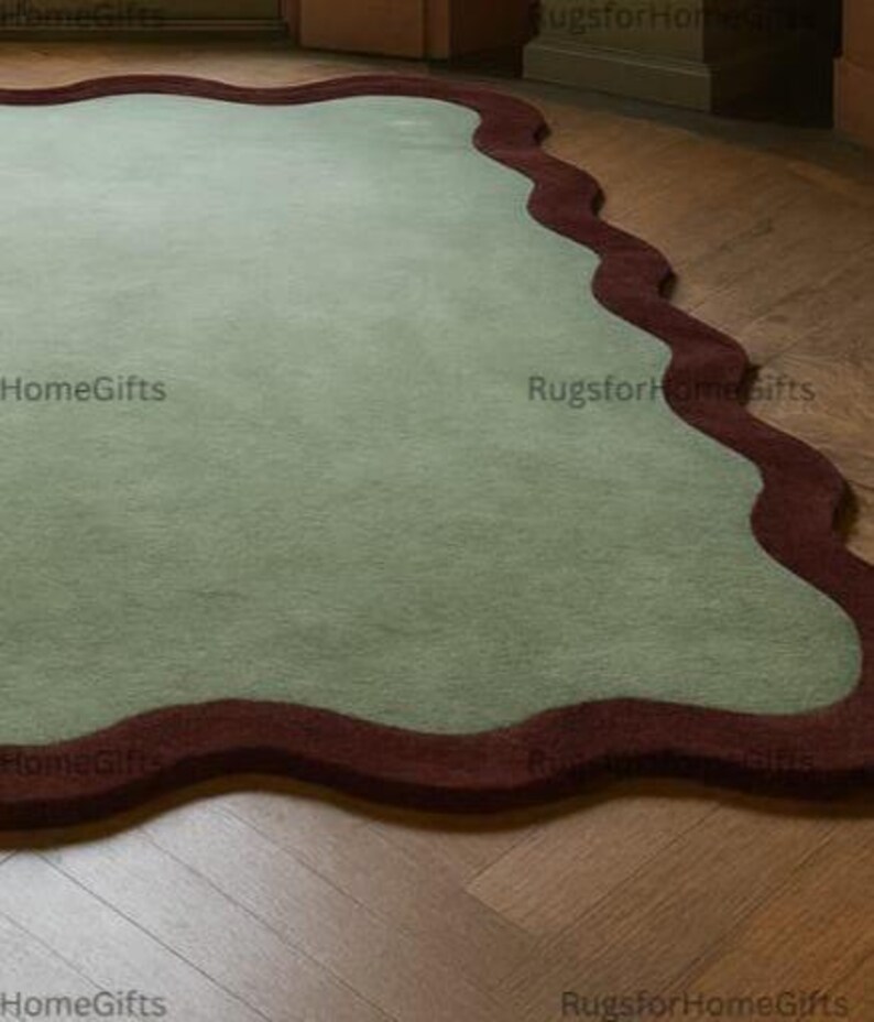May include: A sage green rug with a wavy, dark red border. The rug has an irregular shape. The rug is placed on a wooden floor, adding a touch of colour and texture to the room.
