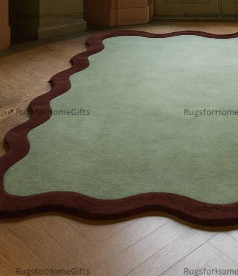 May include: A large, sage green rug with a wavy, dark red border. The rug is placed on a hardwood floor with a herringbone pattern. The rug's unique shape and colour palette add a modern touch to the room.