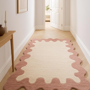 Op de afbeelding: Een rechthoekig vloerkleed met een golvende, onregelmatige rand. Het vloerkleed heeft een lichtbeige midden en een stofroze rand. Het vloerkleed ligt op een lichtgekleurde houten vloer in een gang.