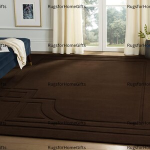 Könnte beinhalten: Ein großer, rechteckiger brauner Teppich mit geometrischem Design. Der Teppich liegt auf einem Holzboden in einem Raum mit einer blauen Chaiselongue, einem gerahmten Bild und einem Fenster mit cremefarbenen Vorhängen.