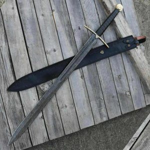 Pode incluir: Uma espada longa e ornamentada com uma lâmina estampada e um cabo preto, repousando sobre uma superfície de madeira. A espada tem uma guarda e um pomo dourados. Uma bainha de couro preta está posicionada por baixo.