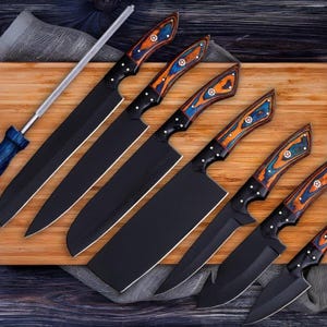May include: A collection of six black kitchen knives with wooden handles, decorated with orange, blue, and white details. The knives are displayed on a wooden board with a sharpening steel. The blades are black.