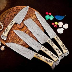 May include: A set of five Damascus steel kitchen knives with cream-colored handles adorned with floral patterns. The blades display a distinctive patterned design. The knives are arranged on a carved wooden surface, with decorative elements nearby.