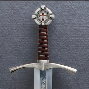 May include: A medieval-style sword with a brown leather-wrapped handle and a silver cross-shaped pommel. The blade is silver and the guard is curved. The pommel features a shield with a cross design.