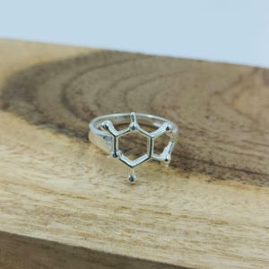 Könnte beinhalten: Ein silberner Ring mit einem Molekülstruktur-Design. Der Ring hat ein geometrisches Muster mit sechseckigen Formen und kleinen Kugeln, vor einem Holzgrund. Der Ring ist ein einzigartiges Schmuckstück.