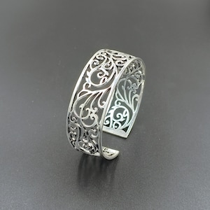 Könnte beinhalten: Ein silberfarbenes Manschettenarmband mit einem komplizierten, durchbrochenen Filigrandesign mit wirbelnden, rankenartigen Mustern. Das Metall hat eine polierte Oberfläche und das Band ist breit mit einer Lücke auf der Rückseite, die ein detailliertes Ausschnittmotiv zeigt.