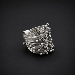 Op de afbeelding: Een zilverkleurige ring opgebouwd uit meerdere dunne, gelaagde draadbanden. Het ontwerp is voorzien van talloze kleine, bolvormige metalen kralen die aan de uiteinden van de draden zijn bevestigd, wat zorgt voor een textuurrijk, gelaagd uiterlijk met een gepolijste afwerking.