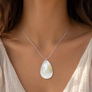 Puede incluir: Un collar de cadena en tono plateado con un gran colgante en forma de lágrima hecho de nácar iridiscente. El colgante muestra una superficie lisa y pulida con variaciones de color naturales en blanco y amarillo dorado sutil.