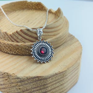 Puede incluir: Un collar con colgante de plata que presenta una piedra preciosa granate roja oscura y redonda, engastada en el centro de un marco estilo sol con rayos metálicos radiantes y detalles de cuentas, suspendido de una fina cadena de plata.