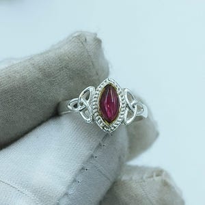 Könnte beinhalten: Silberring mit einem marquiseförmigen, tiefroten Edelstein. Der Ring hat ein keltisches Knotenmuster an den Seiten und einen goldfarbenen Rand um den Edelstein. Der Ring wird auf einer hellgrauen Oberfläche präsentiert.