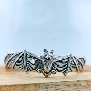 May include: A silver-toned metal cuff bracelet featuring a detailed bat design with outstretched wings. The bat is centered on the band, displaying textured wing patterns and defined facial features in a polished metallic finish.