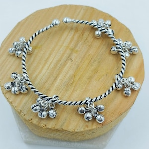 Puede incluir: Una pulsera de metal plateado con una banda trenzada estilo cuerda. La banda circular está adornada con múltiples grupos de pequeñas campanas esféricas espaciadas uniformemente alrededor de toda la circunferencia de la pieza.