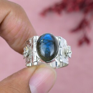 May include: A silver ring with an oval, blue-green gemstone. The ring has a decorative band with floral and geometric details. The gemstone is the focal point, displaying a play of colors. The ring is a piece of jewelry.