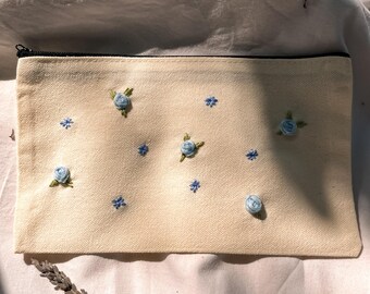 May include: A rectangular, cream-colored canvas pouch with a black zipper. The pouch is adorned with embroidered light blue roses and small blue floral accents. The embroidery is detailed and adds a delicate touch to the accessory.