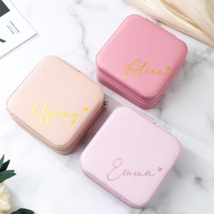 Peut inclure: Trois boîtes à bijoux carrées roses avec des lettres dorées. Les boîtes sont personnalisées avec les noms "Tiffany", "Alice" et "Emma". Chaque boîte a une fermeture éclair et un symbole de cœur à côté du nom.