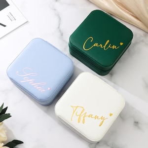 May include: Three square jewelry cases in pastel colors: light blue, green, and white. Each case has a name in gold script: "Sophia", "Carlin", and "Tiffany". The cases are closed and appear to be made of leather or a similar material.