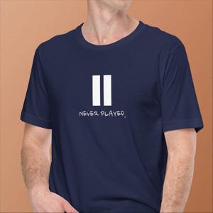 May include: A navy blue t-shirt featuring a white pause symbol and the words "NEVER PLAYED." The short-sleeved shirt is made of a soft fabric. The design is a simple, graphic statement.