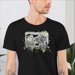 May include: Black t-shirt featuring a distressed camera graphic with white daisies and green leaves. The text "Soft Reset" is below the camera design. The shirt is made of a soft material.