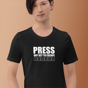 Puede incluir: Camiseta negra con el texto blanco "PRESS ANY KEY TO ESCAPE" y una disposición de teclado de las letras E, P, A, C, S y E. La camiseta es de cuello redondo.