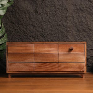 Puede incluir: Un cofre de almacenamiento rectangular de madera con nueve cajones dispuestos en una cuadrícula de tres por tres. El cofre está hecho de madera de color marrón medio con una veta visible. Un cajón en la esquina superior derecha tiene un pomo cuadrado.