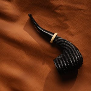 May include: A black tobacco pipe featuring a deeply carved, textured bowl with horizontal ridges. The pipe has a smooth, curved black stem and a small, light-colored circular ring accent where the stem meets the bowl, creating a contrast.