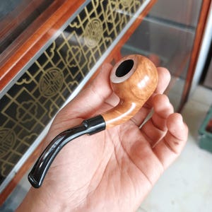 Puede incluir: Una pipa de tabaco curvada, de color marrón claro, con boquilla y caña negros. El cuenco de la pipa tiene un patrón de vetas de madera. La pipa está en la mano de una persona. La longitud de la pipa es de unos 15 cm.