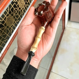 Op de afbeelding: Een handgemaakte tabakspijp met een donkerbruine ketel en een lichte bamboe-stijl steel. De pijp heeft een zwart, getextureerd eindstuk. De ketel heeft een uniek, gesneden ontwerp. De pijp wordt in een hand gehouden.
