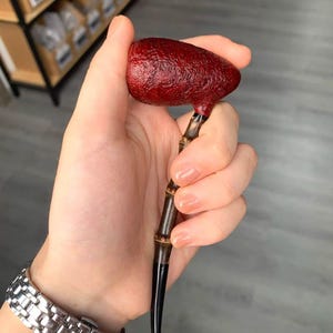 May include: A tobacco pipe featuring a textured, deep red bowl and a bamboo stem. The stem transitions to a black finish. The pipe is held in a hand, highlighting the craftsmanship and the natural materials used in its construction.