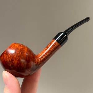 May include: A tobacco pipe featuring a polished, reddish-brown wooden bowl with a visible natural grain pattern. The pipe has a smooth, curved transition into a sleek, black acrylic stem that extends outward, ending in a narrow, tapered mouthpiece.