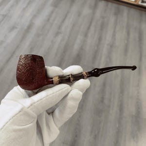 May include: A brown tobacco pipe with a textured bowl and a bamboo-style stem. The pipe has a dark, curved mouthpiece. The bowl is a deep reddish-brown color.