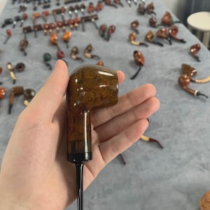 May include: A collection of tobacco pipes, with one held in a hand. The pipes vary in shape and color, with a focus on brown and tan tones. The pipes are made of wood and have a classic design.