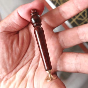 May include: A dark brown wooden wax seal stamp with a brass base. The stamp has a long, slender handle and a decorative top. The brass base is circular and slightly flared. The wood grain is visible.