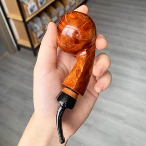 Puede incluir: Una pipa de tabaco artesanal con un cuenco redondeado y una boquilla curvada. La pipa está hecha de madera pulida con un color marrón rojizo intenso. La boquilla y la boquilla son negras, con un anillo de acento marrón claro.