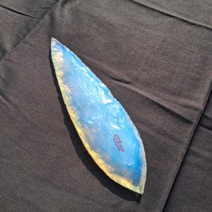 Peut inclure: Un cristal poli et allongé avec une pointe. Le cristal a un aspect translucide et opalescent avec des nuances de bleu et de jaune. Le cristal est posé sur un tissu sombre. Le texte "Miracle Crystal" est visible sur le cristal.