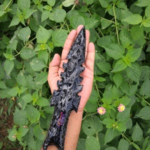 May include: A black obsidian arrowhead with a detailed, textured design. The arrowhead is held in a hand against a backdrop of green foliage. The arrowhead has a pointed tip and a decorative handle.