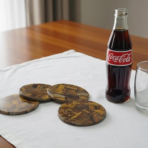 May include: A still life arrangement showing a Coca-Cola bottle with a red and white label, a clear glass with ice, and several oval coasters. The coasters have a brown and gold mosaic pattern. The items are on a white tablecloth.