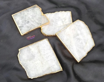May include: Four square, white stone coasters with gold-colored edges are arranged on a black, textured surface. The coasters have a natural, irregular shape and a polished finish. The brand name "Miracle Crystal" is visible.