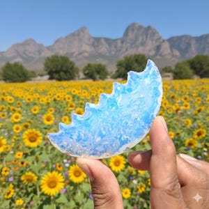 Puede incluir: Un cristal translúcido azul claro en forma de media luna con un borde dentado. El cristal se sostiene frente a un campo de girasoles y montañas bajo un cielo azul claro.