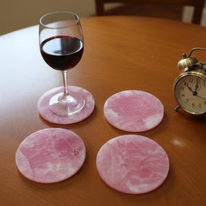May include: A glass of red wine rests on a pink stone coaster, with three more coasters nearby. A vintage brass alarm clock is also on the wooden table. The coasters have a marbled appearance.